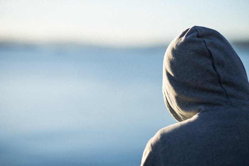 Anonymisert bilde av en person med ryggen til og hette trukket over hodet. Ser ut mot havet.
